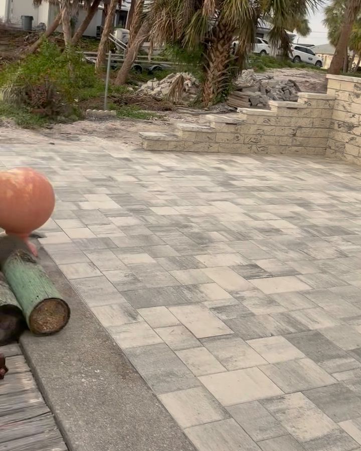 Large gray and tan paver patio with a stone retaining wall and palm trees